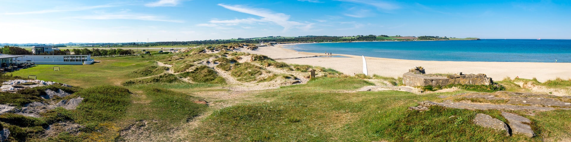 A coastal landscape with a sandy beach, grassy dunes, and a clear blue sky.