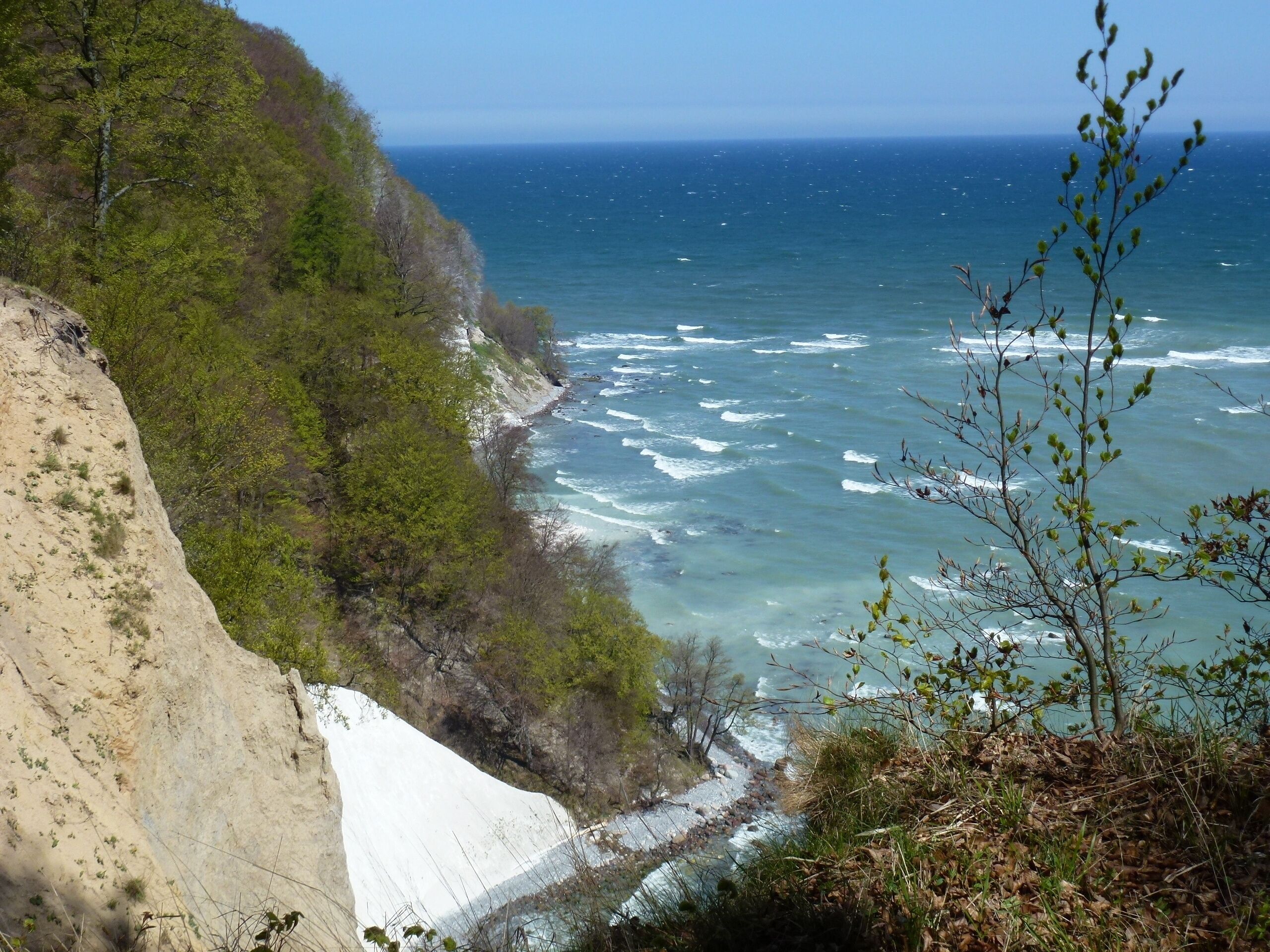 A coastal cliff with waves crashing against it, a clear sky, and a distant view of the ocean.