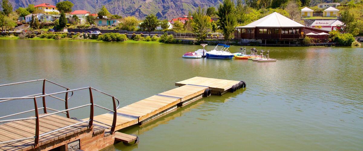 A wooden dock extending into a calm lake with boats docked, surrounded by lush greenery and a mountain range in the background.