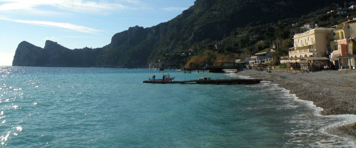 A coastal scene with a clear blue sea, boats on the water, a rocky mountain in the background, and buildings along the shore.
