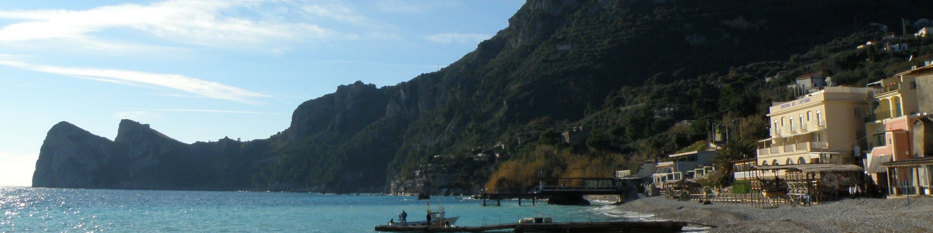A coastal scene with a clear blue sea, boats on the water, a rocky mountain in the background, and buildings along the shore.