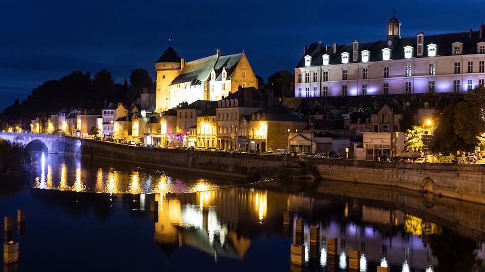 A nighttime river scene with a stone bridge, illuminated buildings, and a castle-like structure.