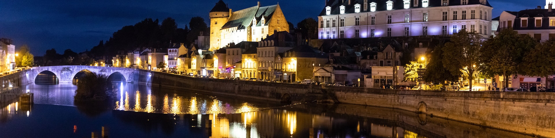 Une scène nocturne au bord d’une rivière, avec un pont de pierre, des bâtiments illuminés et une structure qui ressemble à un château.