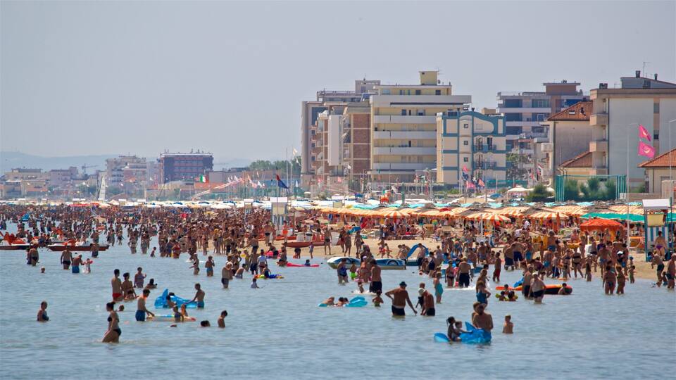 A crowded beach with people swimming and sunbathing, umbrellas, and buildings in the background.
