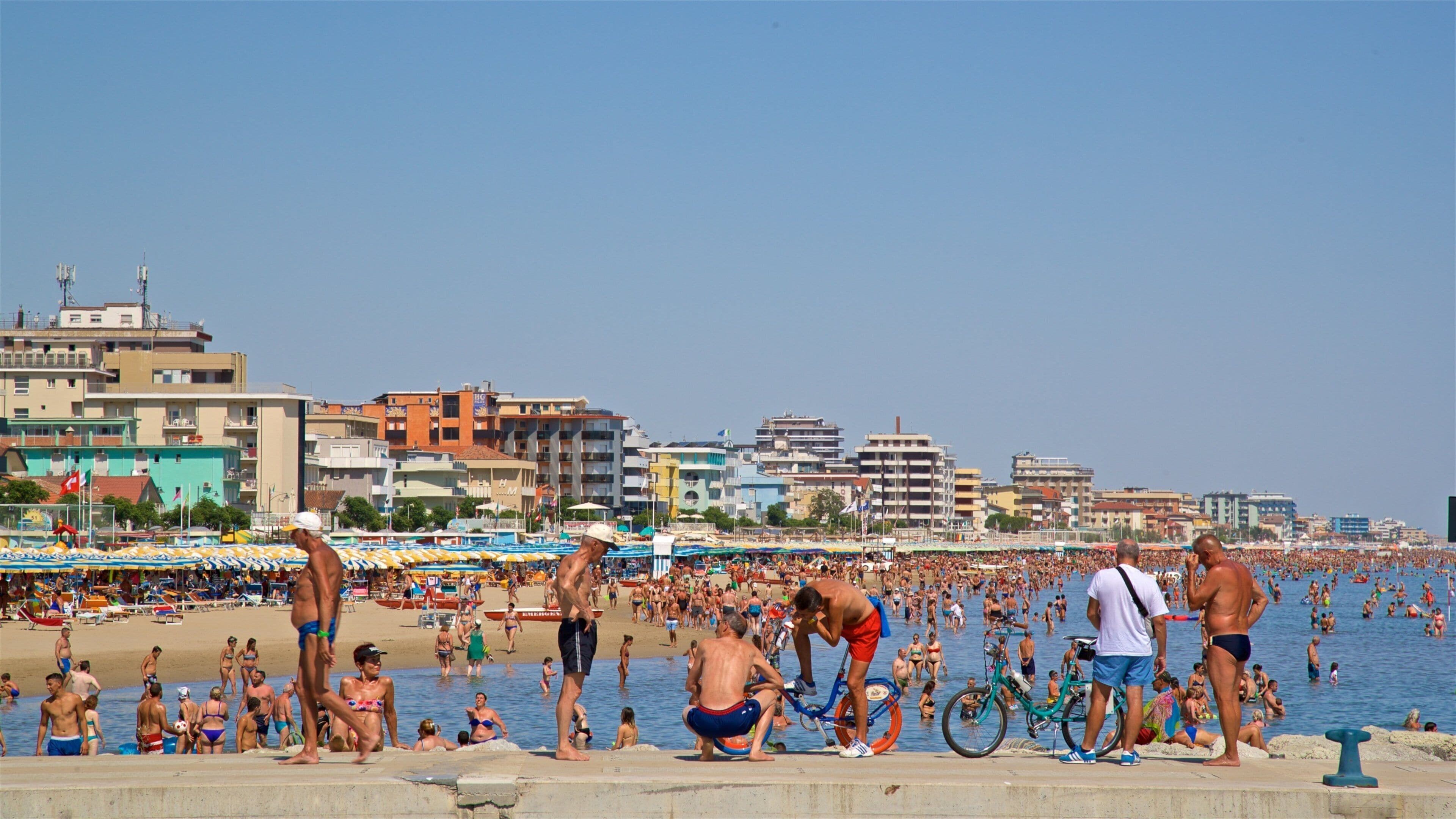 A crowded beach with people sunbathing and swimming, surrounded by colorful buildings and a clear blue sky.