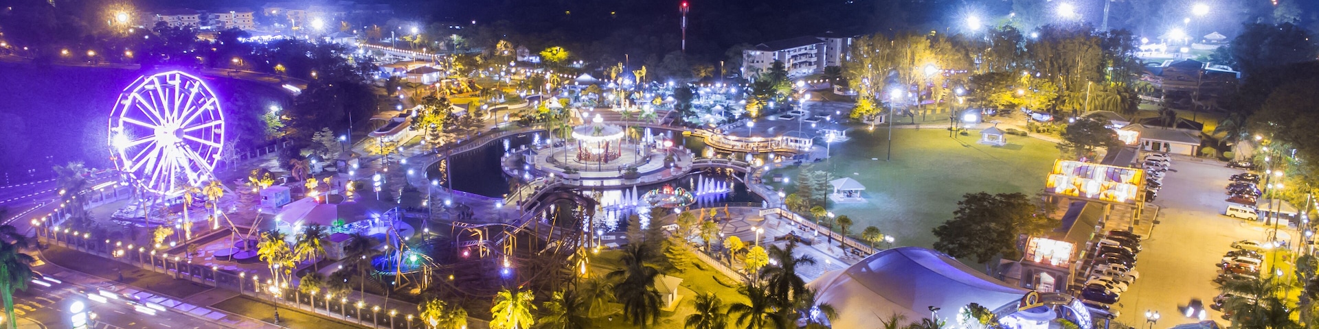 Um parque de diversões iluminado à noite, com uma grande roda-gigante, fontes de água e caminhos bem iluminados.