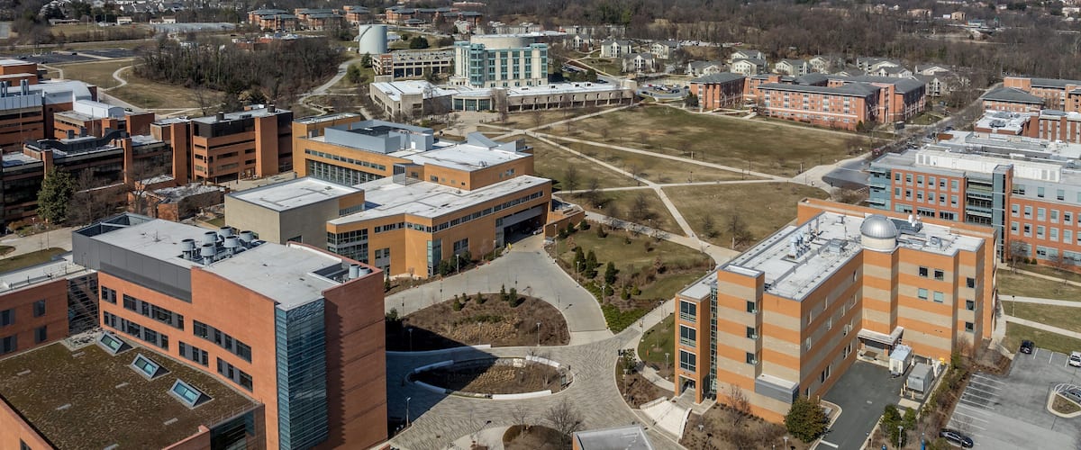 An aerial view of a university campus with modern buildings, green spaces, and a clear blue sky.