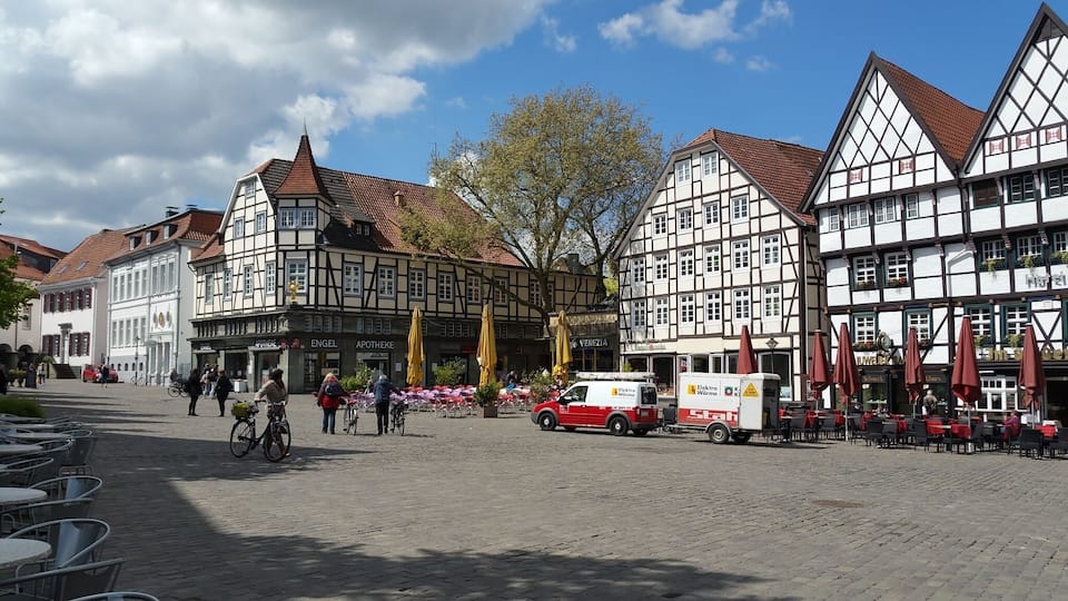 A cobblestone street lined with traditional half-timbered buildings, a red delivery truck, and outdoor seating with umbrellas.