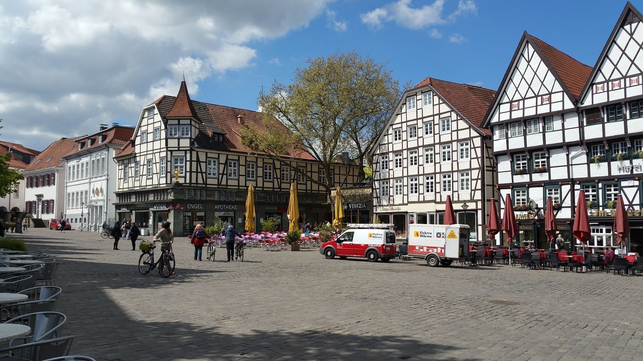 A cobblestone street lined with traditional half-timbered buildings, a red delivery truck, and outdoor seating with umbrellas.