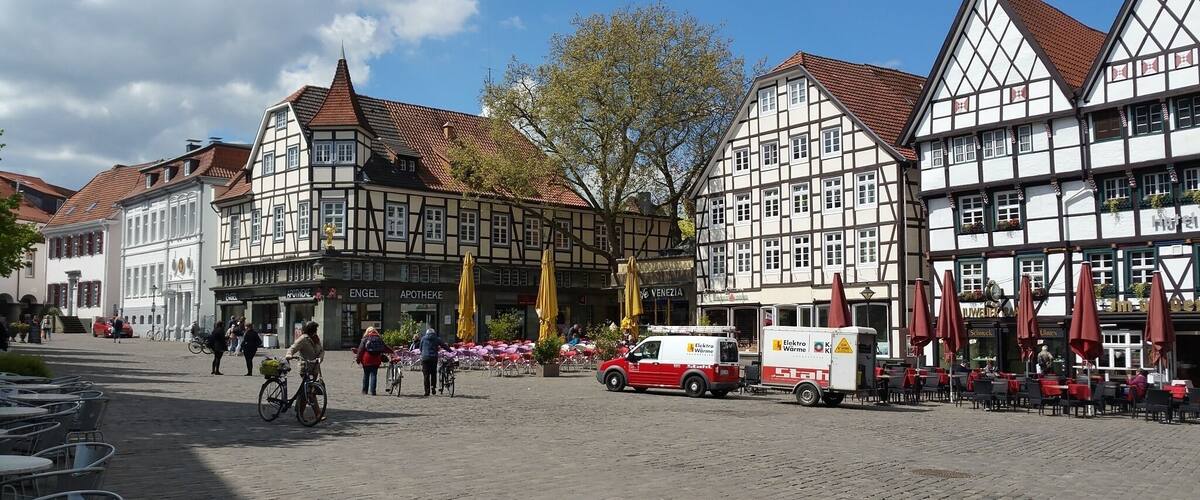 A cobblestone street lined with traditional half-timbered buildings, a red delivery truck, and outdoor seating with umbrellas.