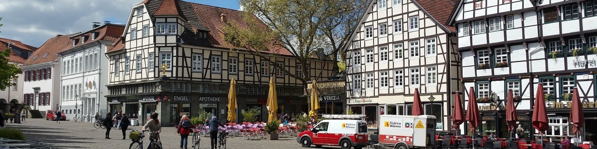 A cobblestone street lined with traditional half-timbered buildings, a red delivery truck, and outdoor seating with umbrellas.