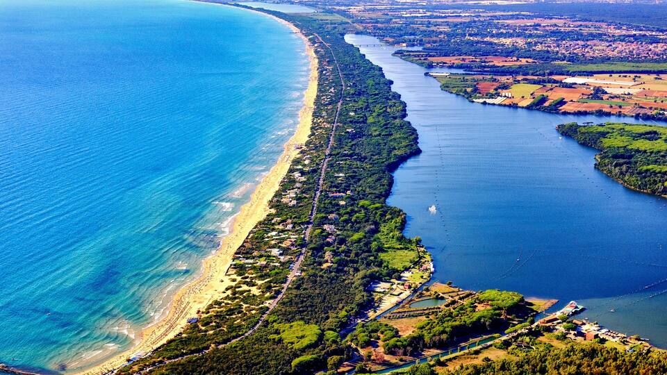An aerial view of a coastal area with a long sandy beach, dense green vegetation, and a river or bay running parallel to the shore.