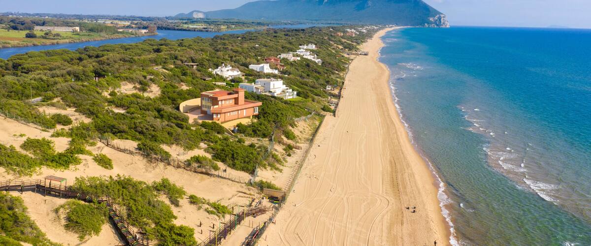 A coastal landscape with a sandy beach, a mountain in the background, and scattered buildings.