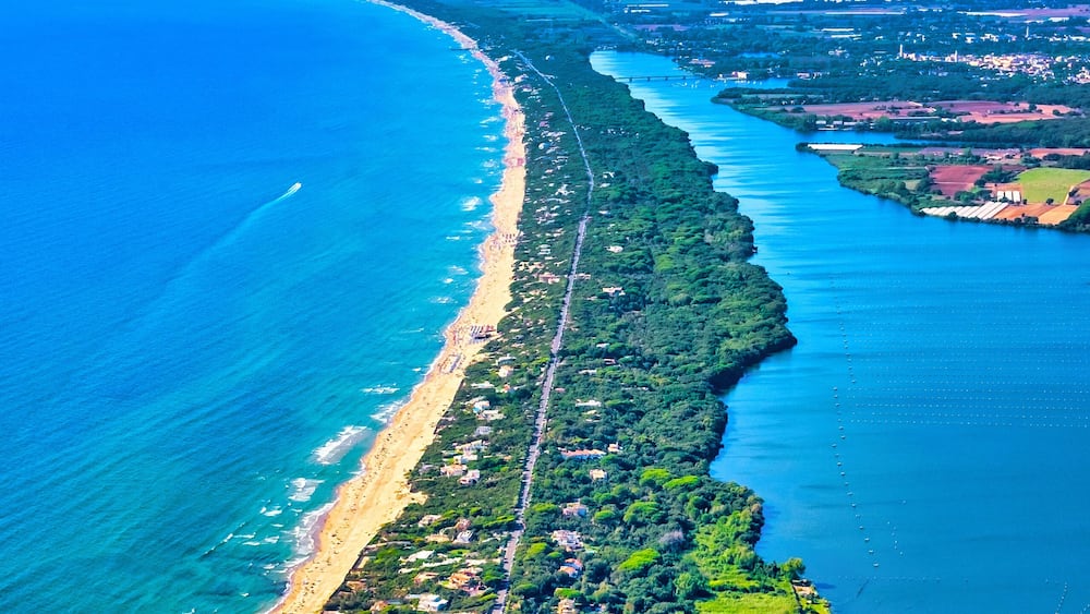 A coastal landscape with a long sandy beach, dense greenery, and a clear blue sea.