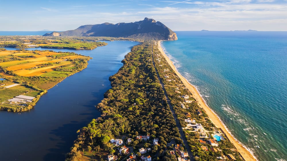 An aerial view of a coastal town with a long tree-lined promenade, a beach, and a mountain in the distance.