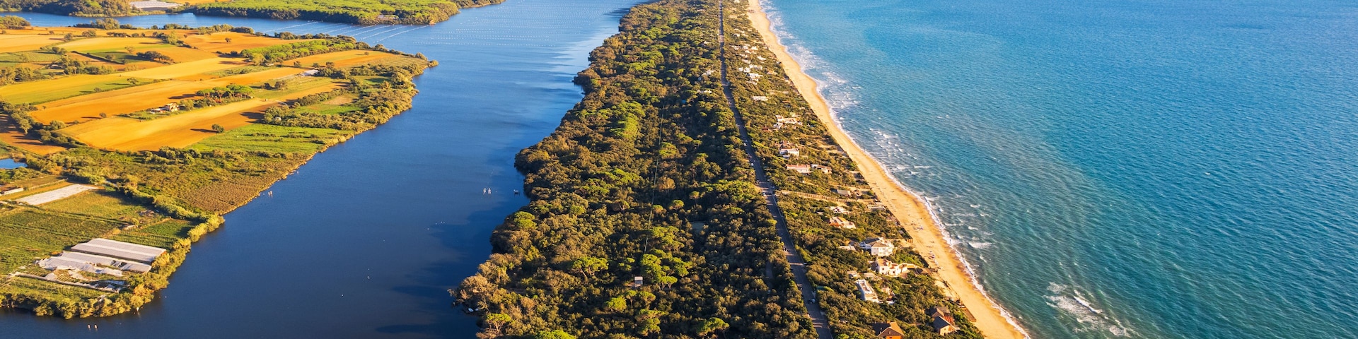 Vista aérea de un pueblo costero con un largo paseo arbolado, una playa y una montaña al fondo.