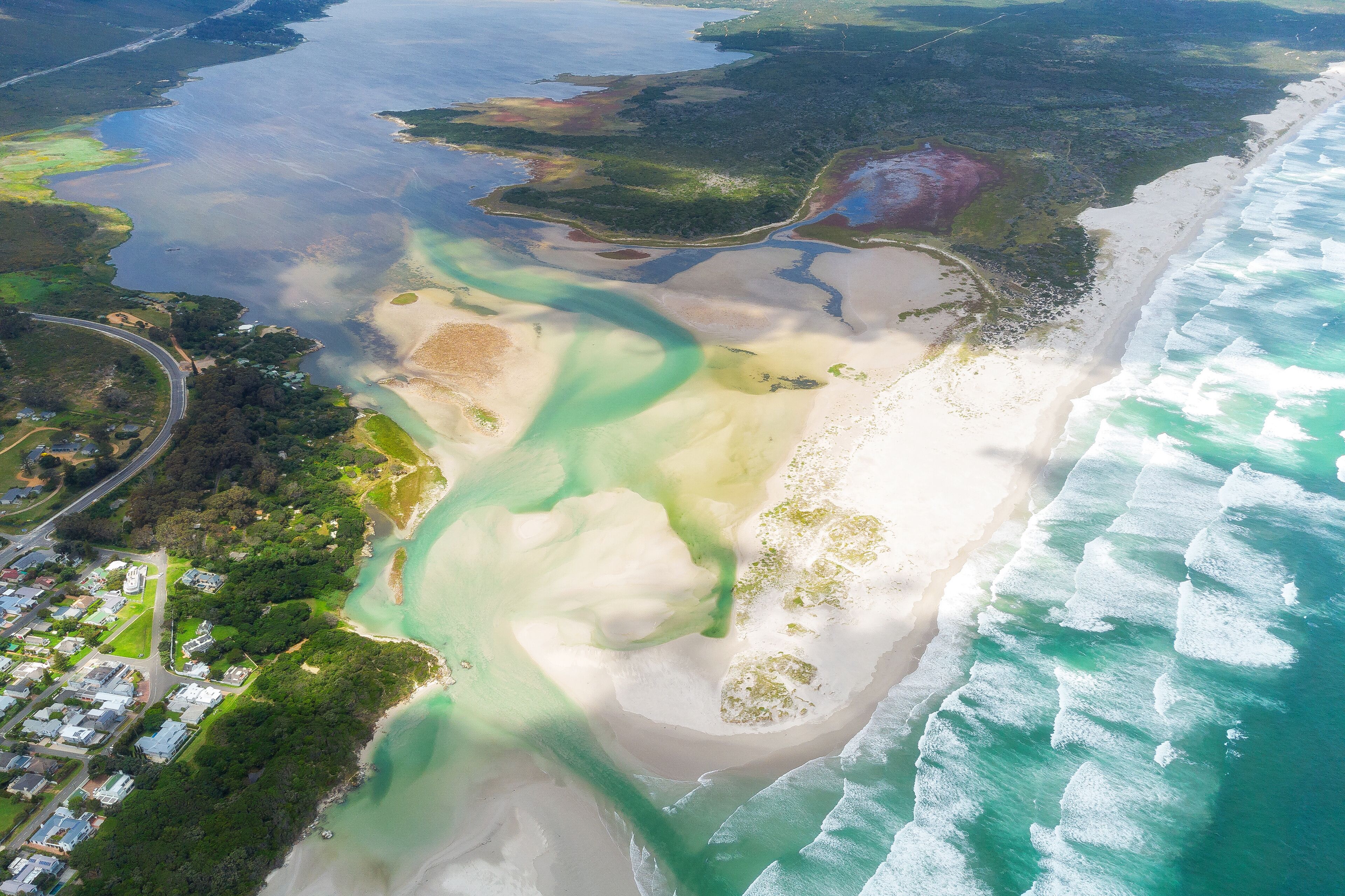 An aerial view of a coastal area with a beach, clear blue waters, and a mix of green vegetation and urban development.