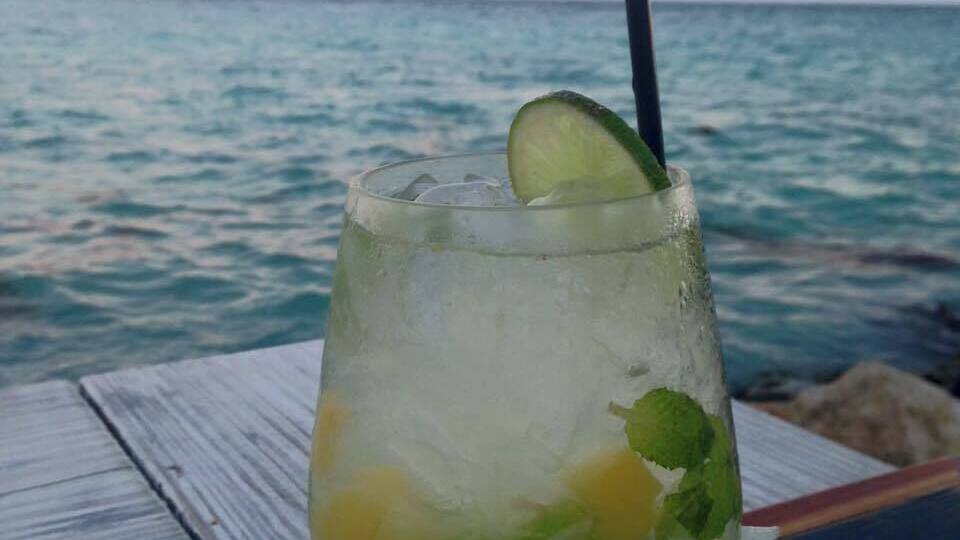 Un verre de boisson agrémenté de citron vert et de menthe, posé sur une table en bois au bord de la mer, au coucher du soleil.