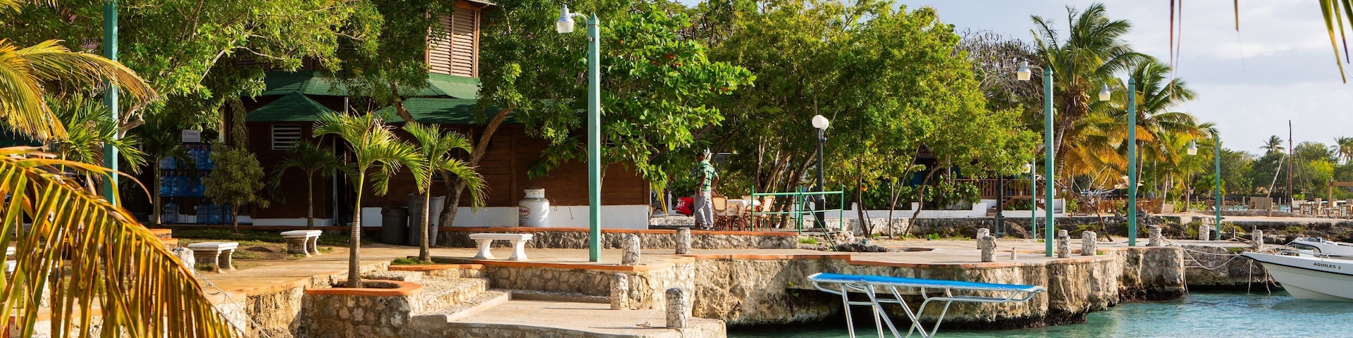 A marina with a white boat named 'Achina' docked, surrounded by palm trees and a building with a green roof.