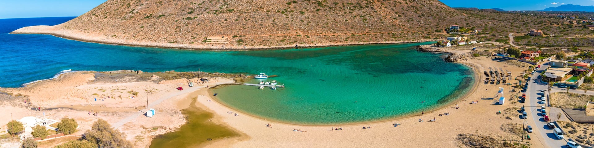 A coastal landscape with a prominent rocky hill, a clear blue sea, a sandy beach, and a small boat docked near the shore.