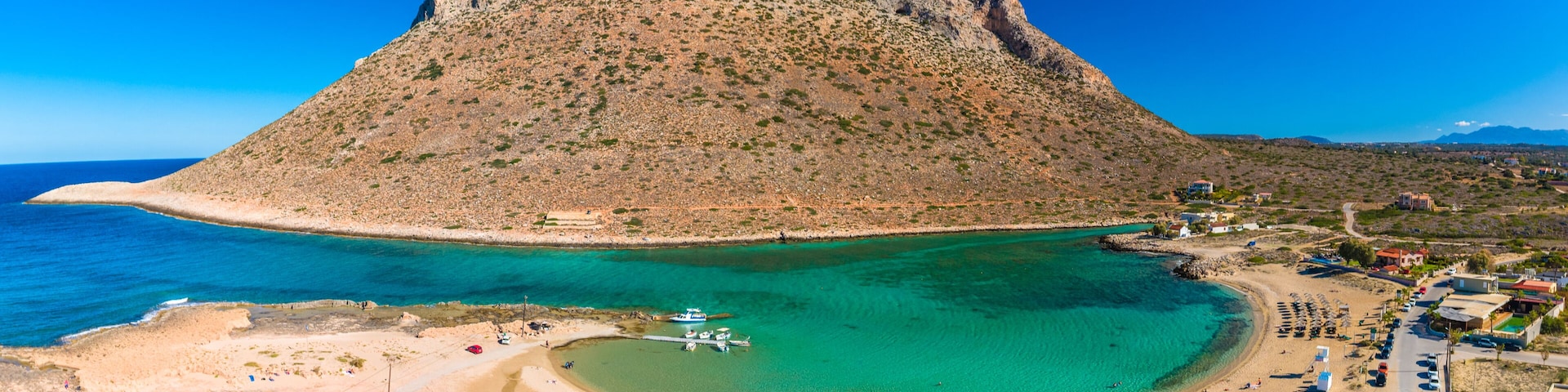 A coastal landscape with a prominent rocky hill, a clear blue sea, a sandy beach, and a small boat docked near the shore.