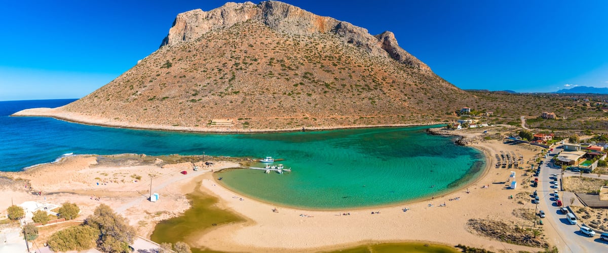 A coastal landscape with a prominent rocky hill, clear turquoise waters, and a beach with parked cars and buildings.