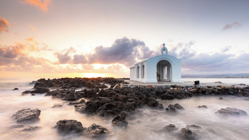 Eine weiße Kirche an einer felsigen Küste mit Sonnenuntergang im Hintergrund.