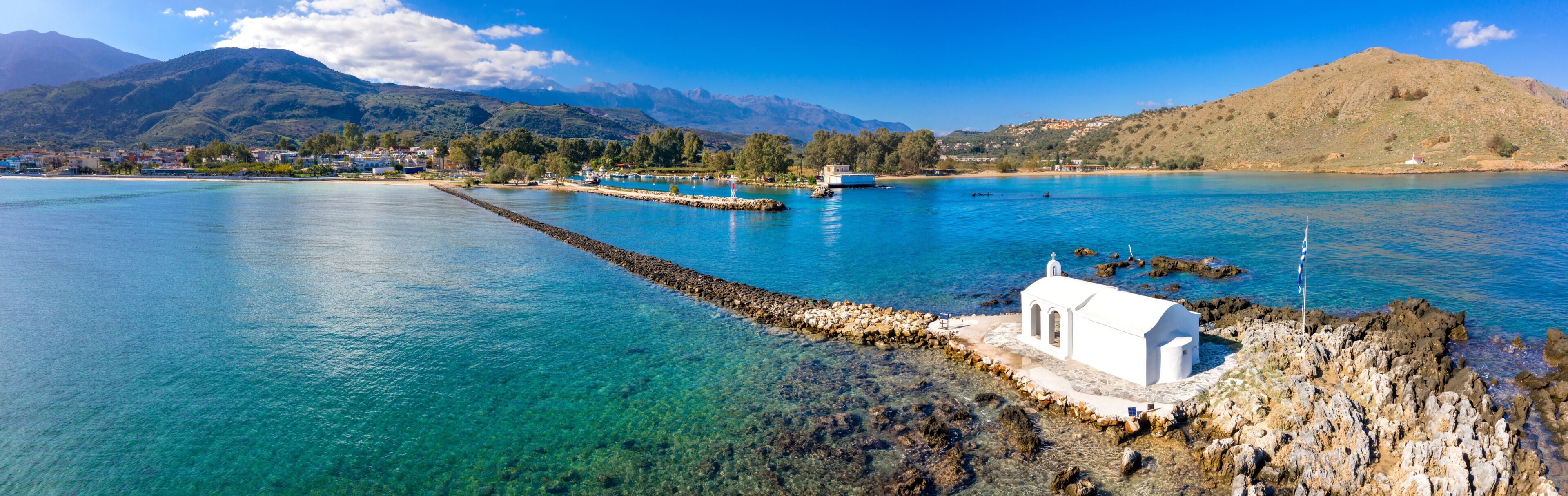 A coastal town with clear blue waters, a white church, and mountains in the background.