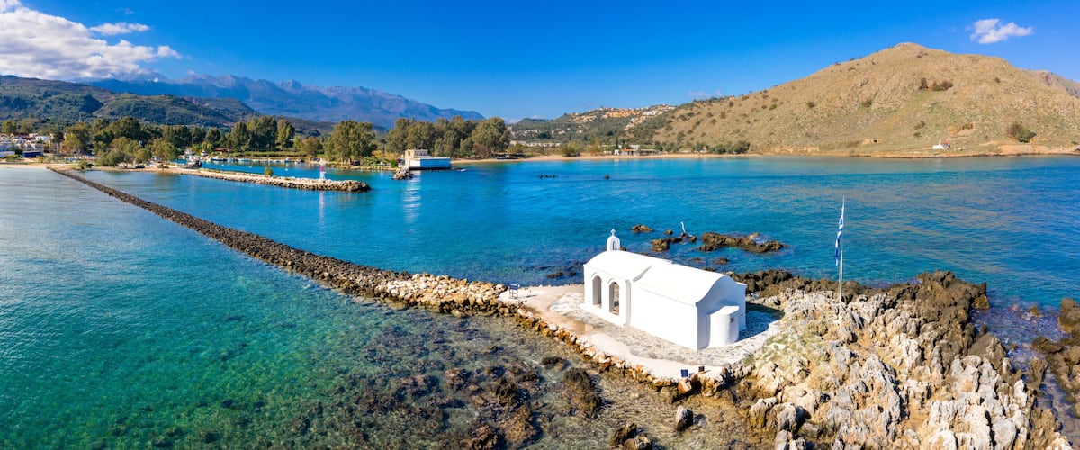 A coastal town with clear blue waters, a white church, and mountains in the background.
