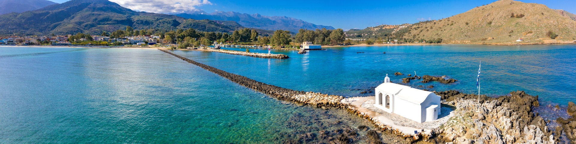 A coastal town with clear blue waters, a white church, and mountains in the background.