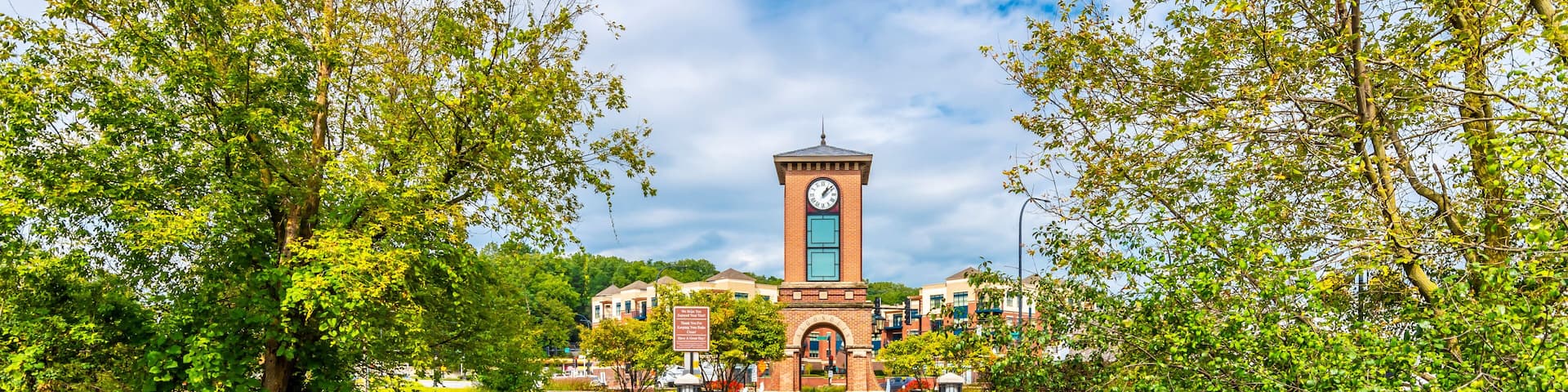 A wooden bridge leading to a clock tower surrounded by lush greenery and a river.