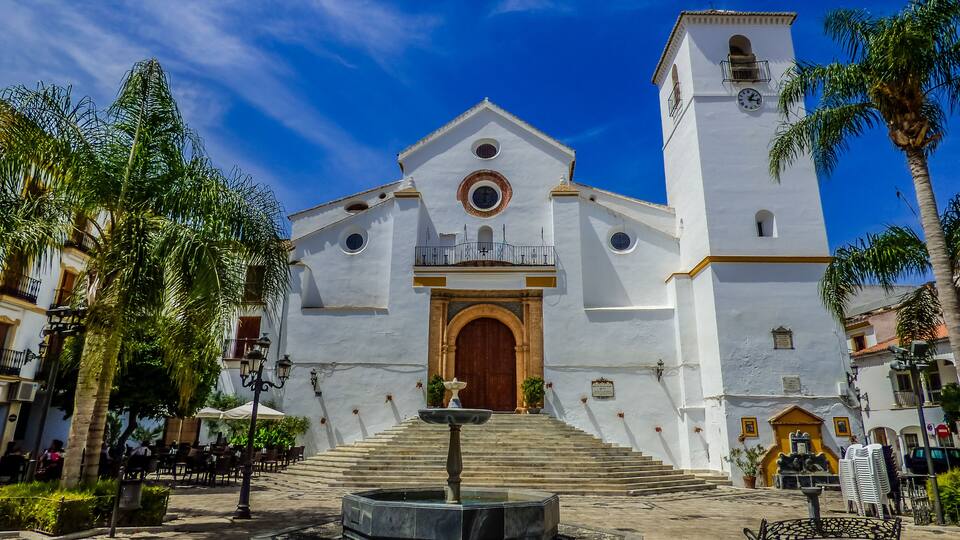 A white church with a clock tower, a fountain, and palm trees.