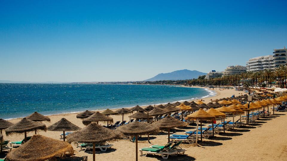 A beach with thatched umbrellas, sun loungers, and a clear blue sea.