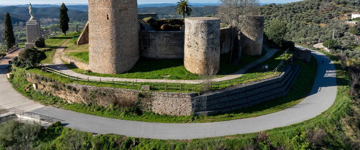 A historic stone fortress with multiple towers, surrounded by a well-maintained garden and a winding road.