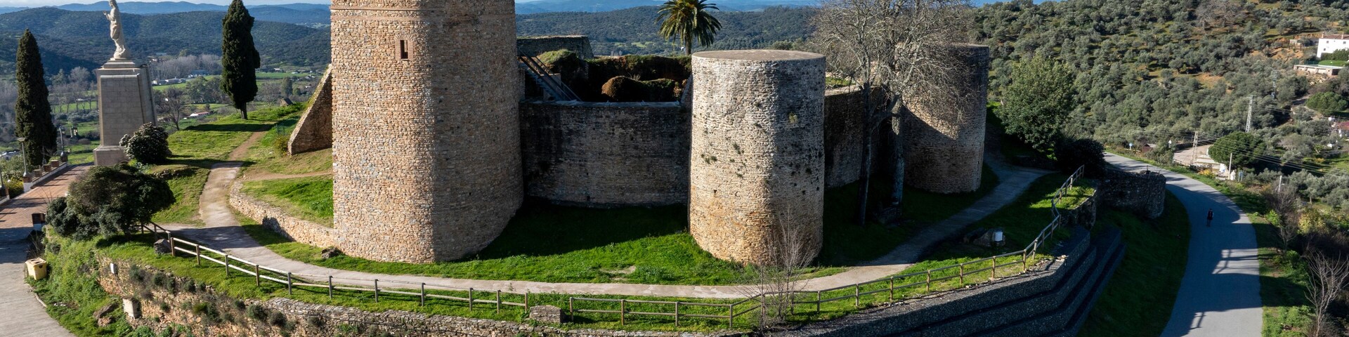 A historic stone fortress with multiple towers, surrounded by a well-maintained garden and a winding road.