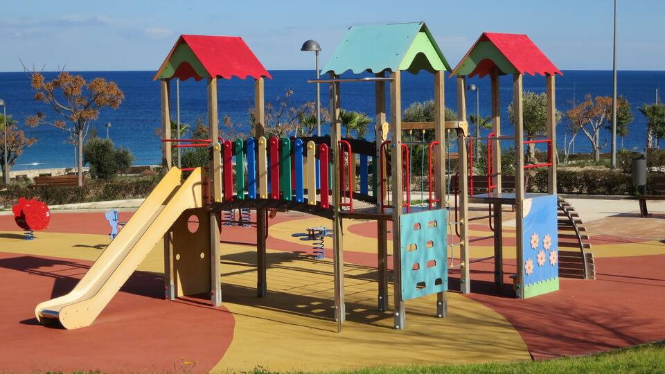 A playground with a slide and climbing structures, set against a backdrop of the sea and clear sky.