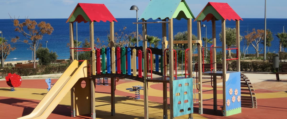 A playground with a slide and climbing structures, set against a backdrop of the sea and clear sky.