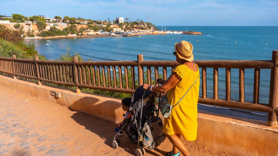 A coastal landscape with a wooden railing, a clear blue sea, and a distant shoreline with buildings.