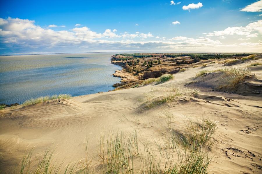 A coastal landscape with sand dunes, grassy areas, and a clear view of the ocean.