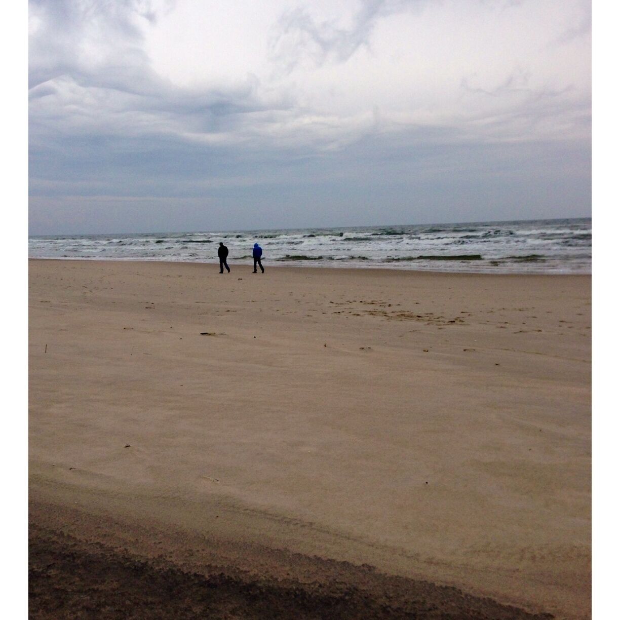 A beach with two people walking along the shore under a cloudy sky.