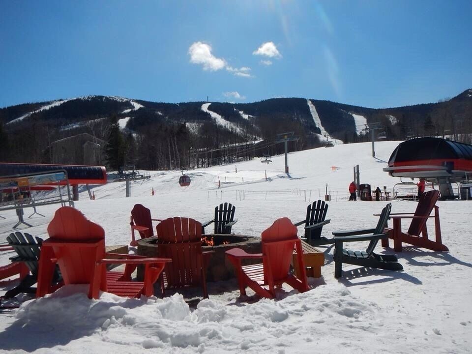 A snow-covered outdoor seating area with red Adirondack chairs and a fire pit, overlooking a ski slope and a mountain range.