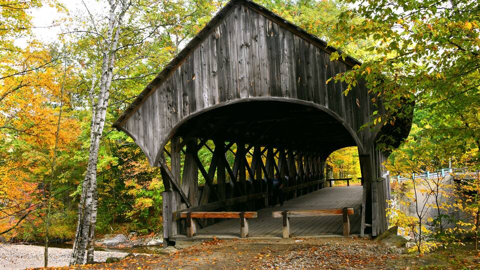 A wooden covered bridge in a forest with autumn leaves.