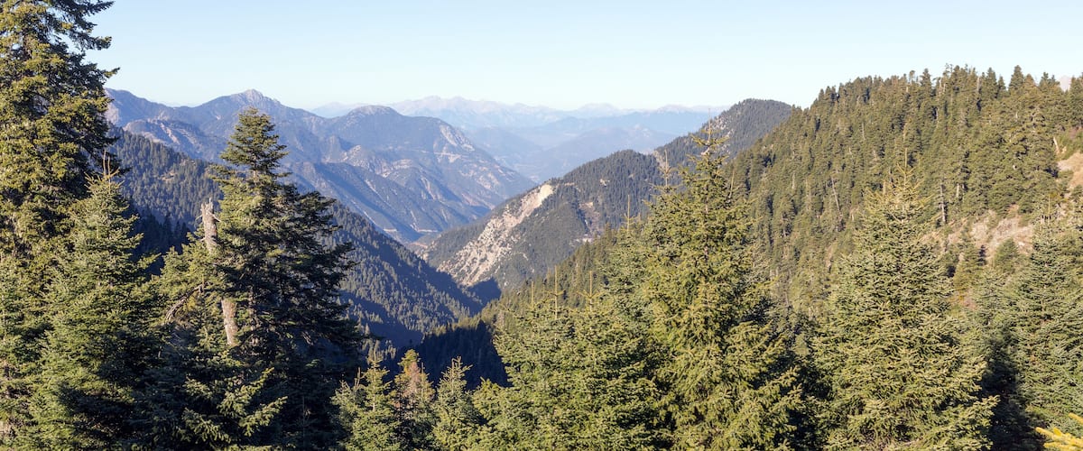 A mountainous landscape with dense forests and distant mountain peaks under a clear sky.