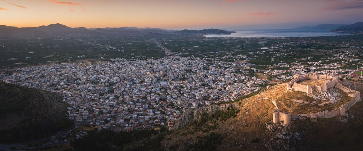 A cityscape with a prominent castle on a hill, surrounded by a mix of urban and rural areas, and a coastline in the distance.