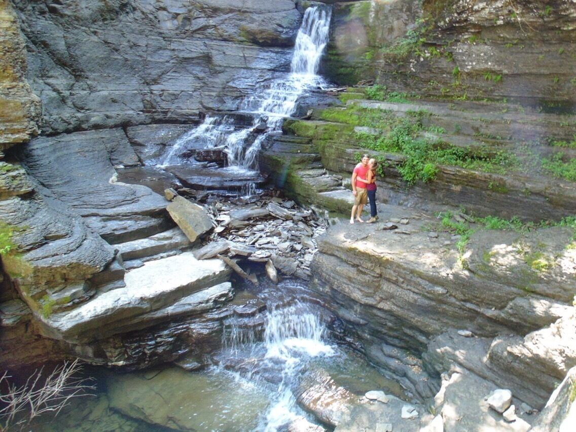 A waterfall cascading over layered rock formations with a person standing on the right side.