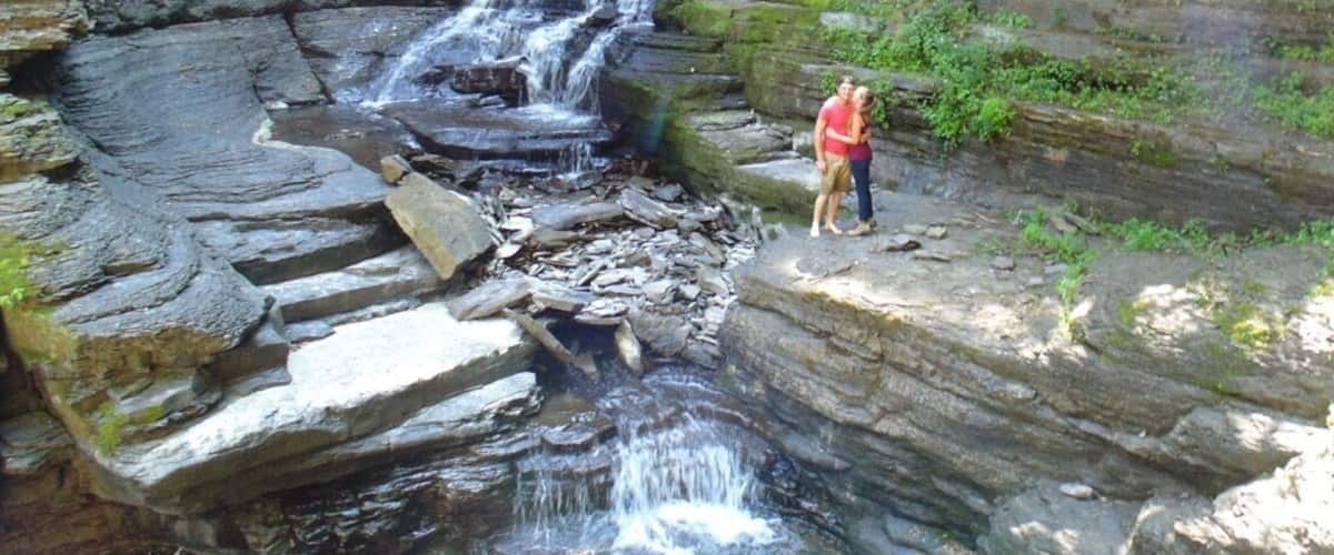 A waterfall cascading over layered rock formations with a person standing on the right side.