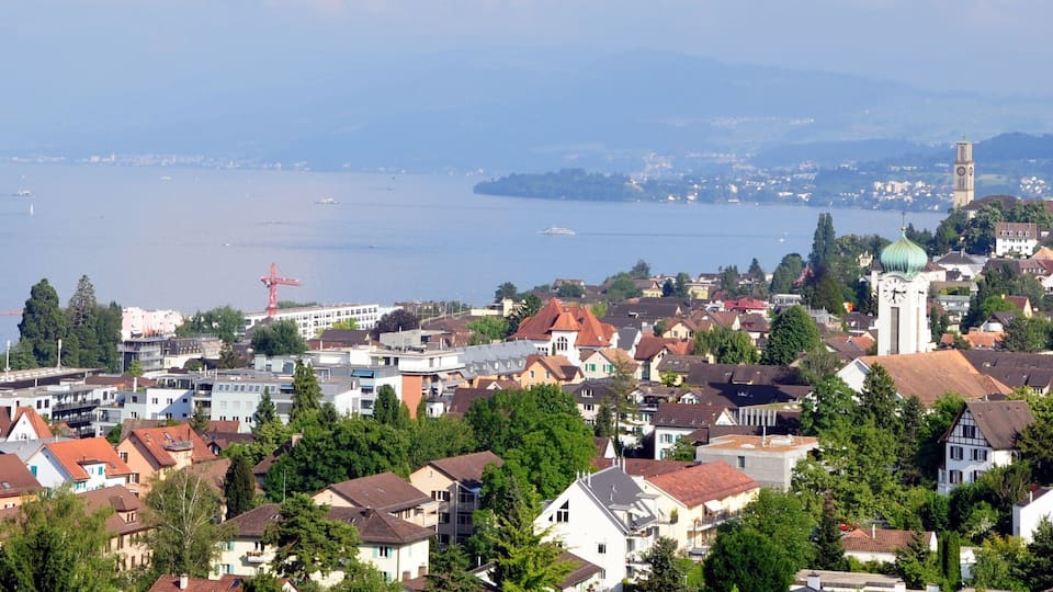 A lakeside town with a prominent clock tower, surrounded by residential buildings and greenery.