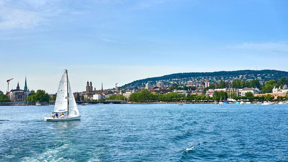 A sailboat on a river with a cityscape and a hill in the background.