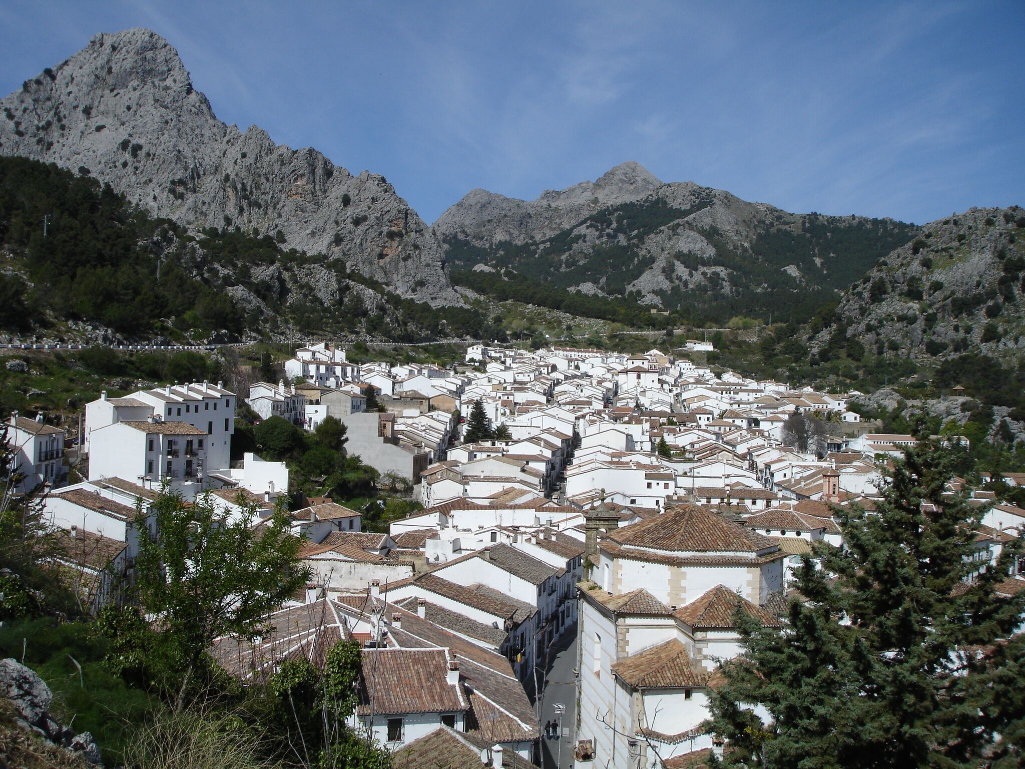 A coastal town with white buildings and terracotta roofs, surrounded by rocky mountains and greenery.