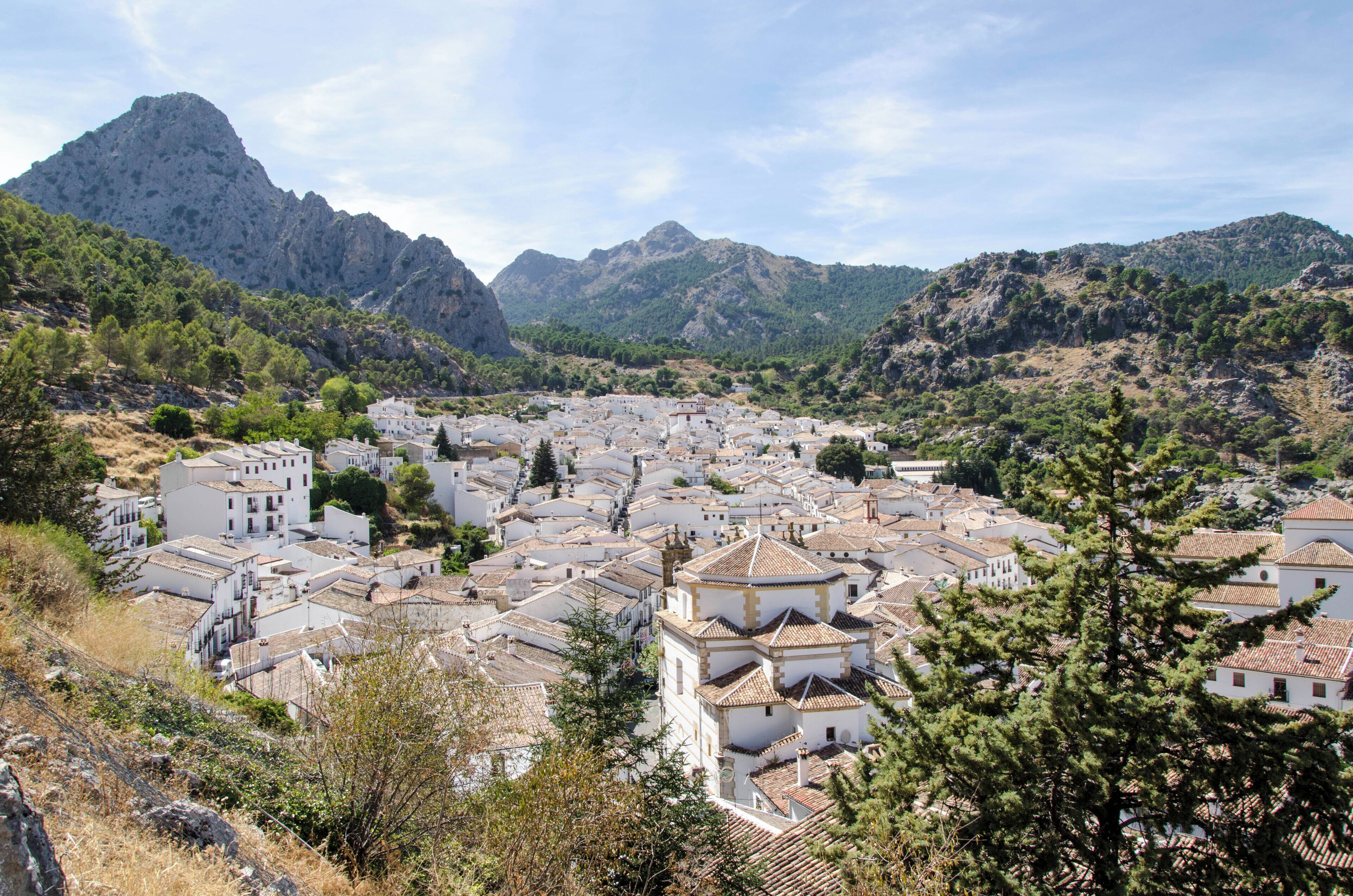 A mountainous town with white buildings and terracotta roofs, surrounded by lush green hills and a prominent rocky peak.
